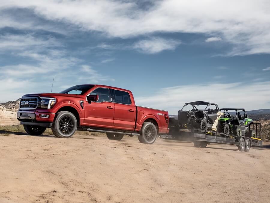 The image shows a red Ford F-150 pickup truck towing a trailer loaded with two off-road vehicles (UTVs). The setting is a dusty trail in a desert-like environment, showcasing the truck’s towing capability and rugged terrain adaptability. The scene captures a sense of adventure and the practical use of the truck in outdoor recreational activities.